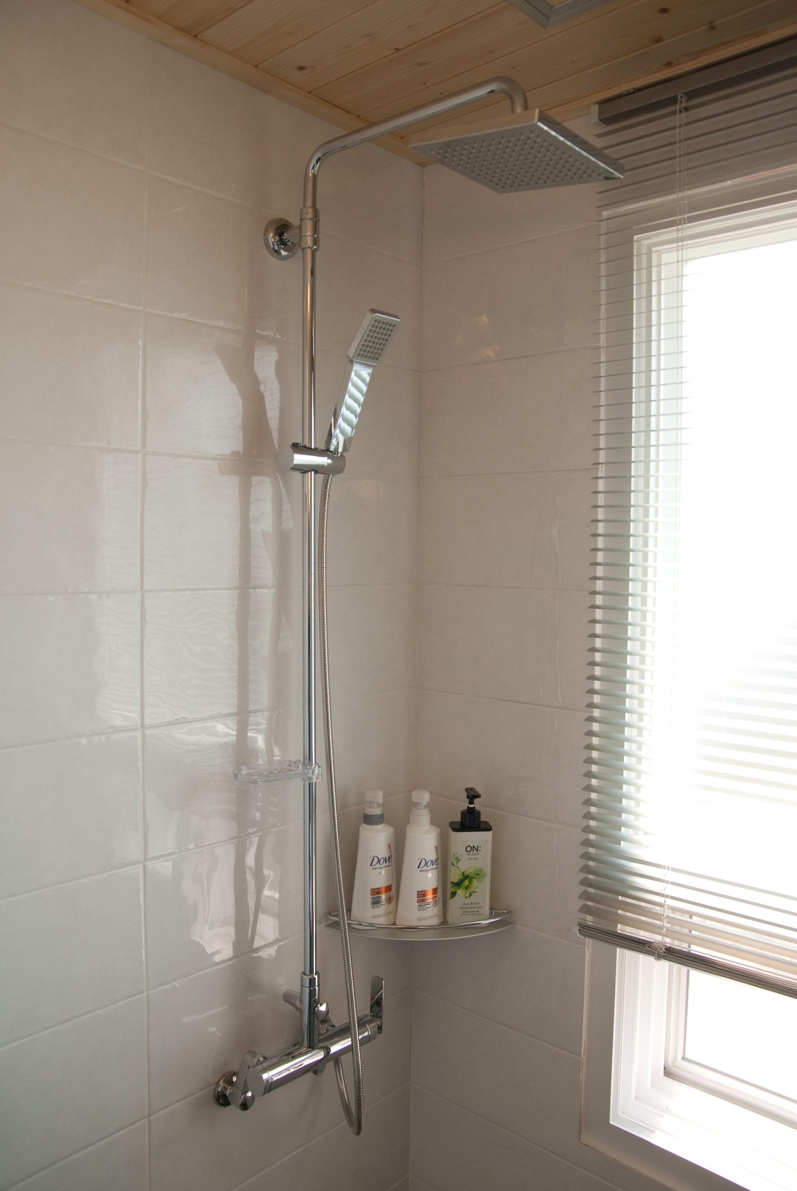 Ducha con cabezal de ducha de mano y grifo empotrado en la pared. Hay dos botellas en un estante debajo de la ducha.