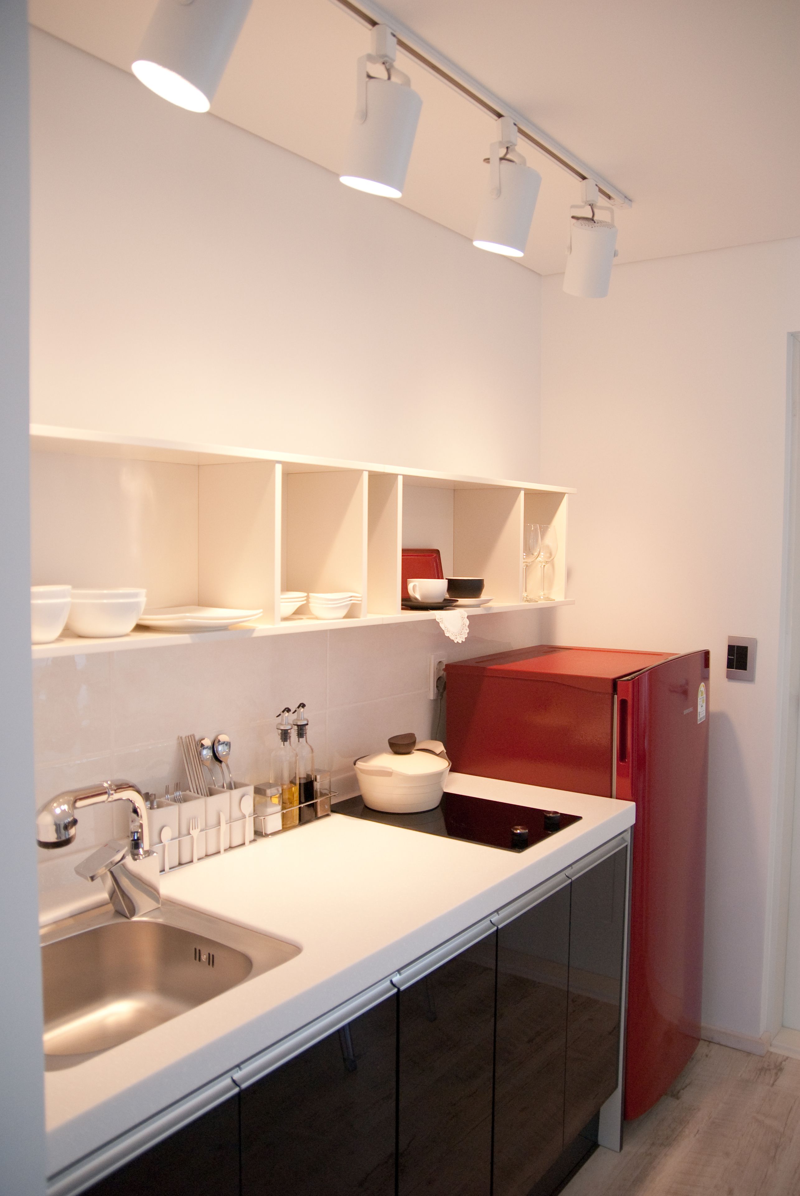 Uma cozinha moderna com bancadas brancas, uma pia de aço inoxidável, uma geladeira vermelha e iluminação embutida.
