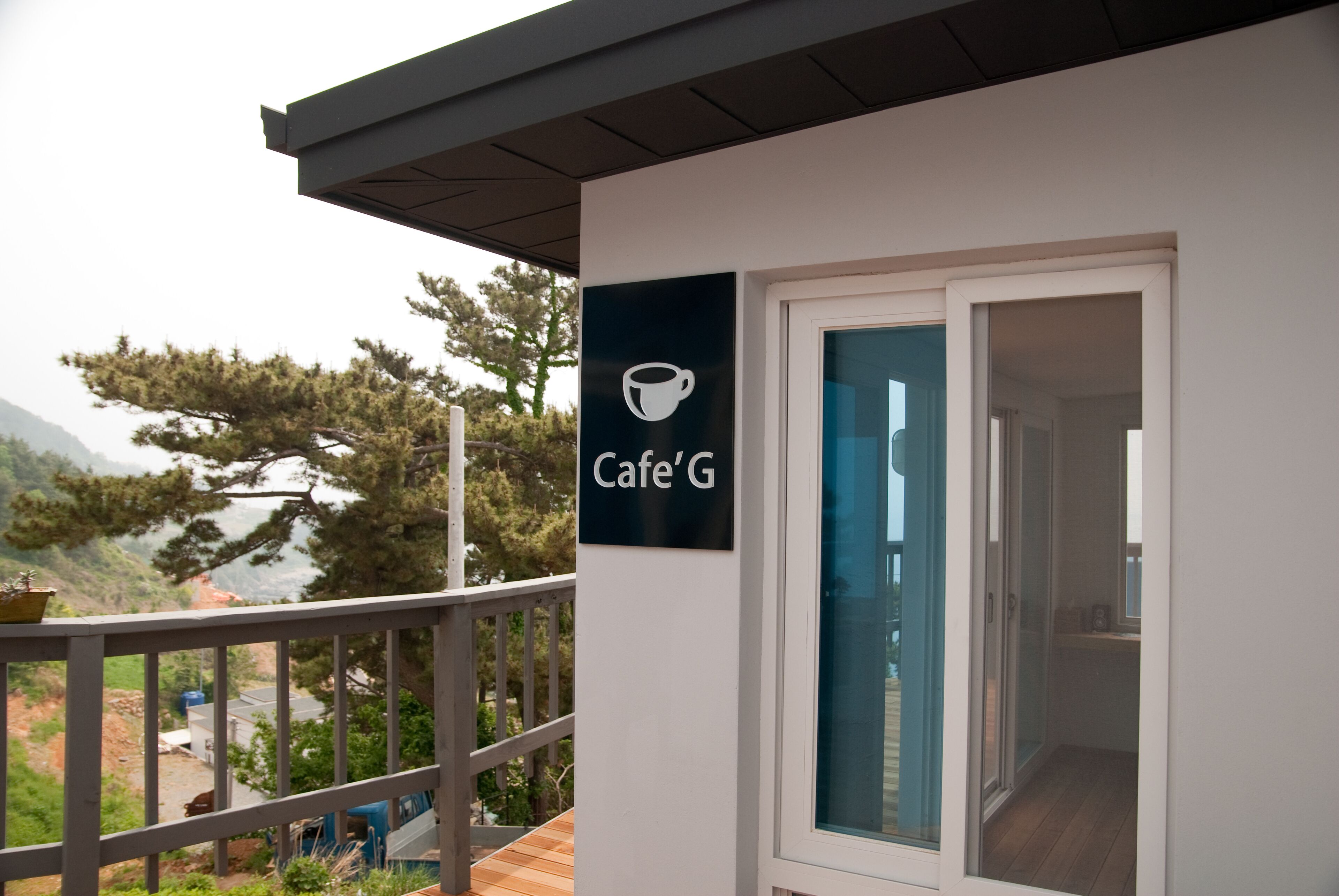Un edificio moderno con un cartel que dice "Cafe'G" en el exterior, un balcón con barandilla y vistas a árboles y montañas al fondo.