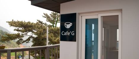 Un edificio moderno con un cartel que dice "Cafe'G" en el exterior, un balcón con barandilla y vistas a árboles y montañas al fondo.