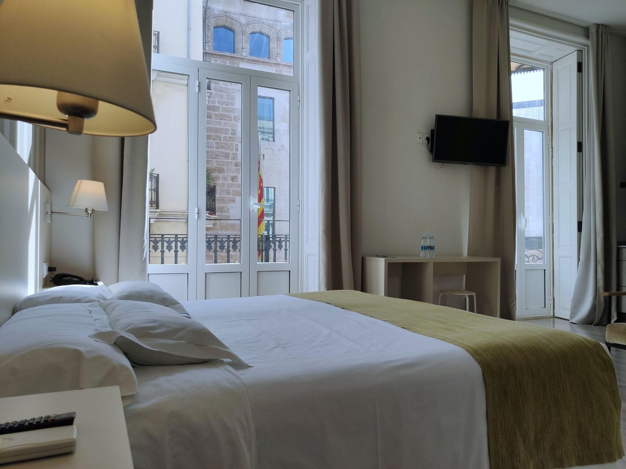 Ein Hotelzimmer mit Bett, Schreibtisch, Fernseher und Fenster mit Blick auf ein Gebäude.