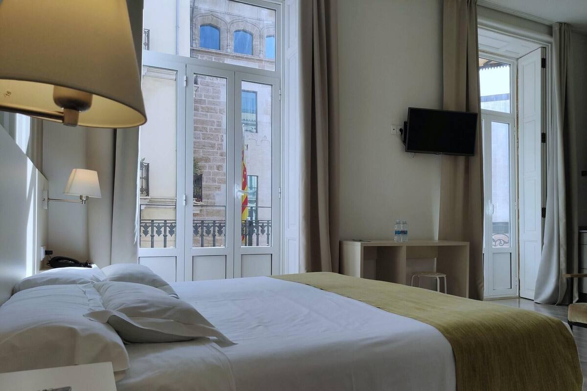 A hotel room with a bed, a desk, a TV, and a window with a view of a building.