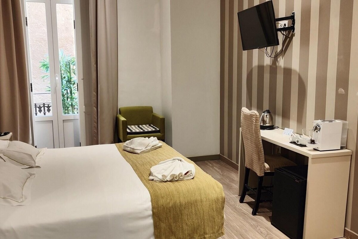 Ein Hotelzimmer mit einem Bett, einem Schreibtisch mit Computer, einem Sessel und einem an der Wand befestigten Fernseher.