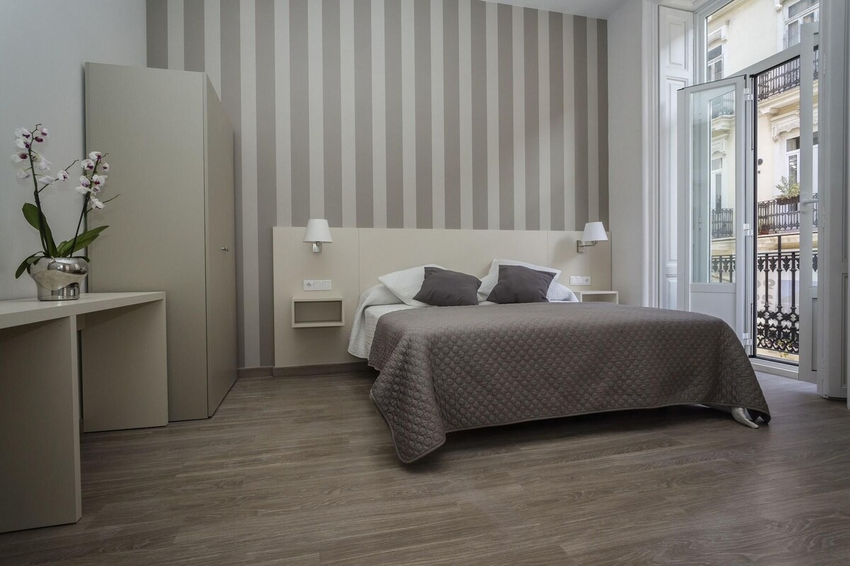 Ein modernes Schlafzimmer mit Bett, Nachttischen, einer Lampe und einem Blumenstrauß, mit Blick auf einen Balkon.