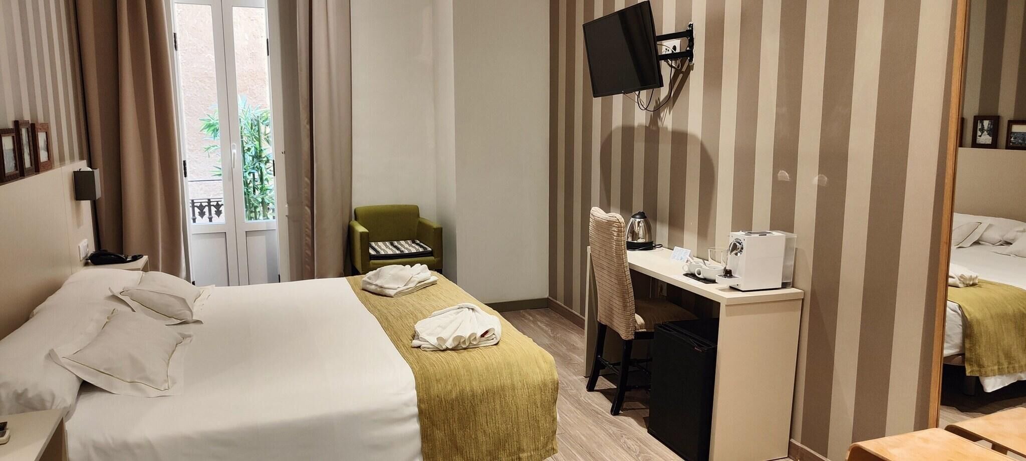 Ein Hotelzimmer mit einem Bett, einem Schreibtisch mit Computer, einem Sessel und einem an der Wand befestigten Fernseher.