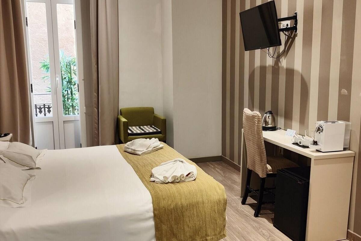 A hotel room with a bed, a desk with a computer, a chair, and a TV mounted on the wall.