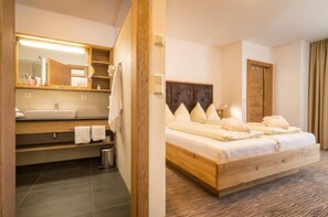 A hotel room with a wooden bed, a large mirror, a sink, and a small trash bin. - Almhotel Karnten (Hermagor-Pressegger See)