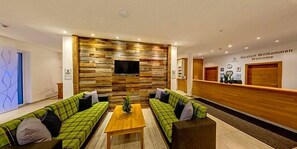A modern lobby with a reception desk, two green plaid sofas, a wooden coffee table, and a wall-mounted TV. - Almhotel Karnten (Hermagor-Pressegger See)