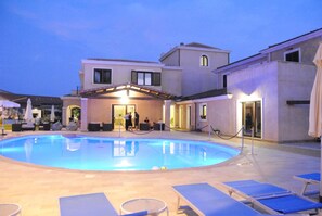 A swimming pool with lounge chairs and umbrellas, surrounded by buildings with a covered patio area. - La Perla (Tortoli)