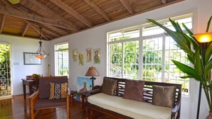 A spacious living area with a wooden ceiling, a sofa, a dining table, and a large window with a view of greenery. - Tranquility Villa (Port Antonio)
