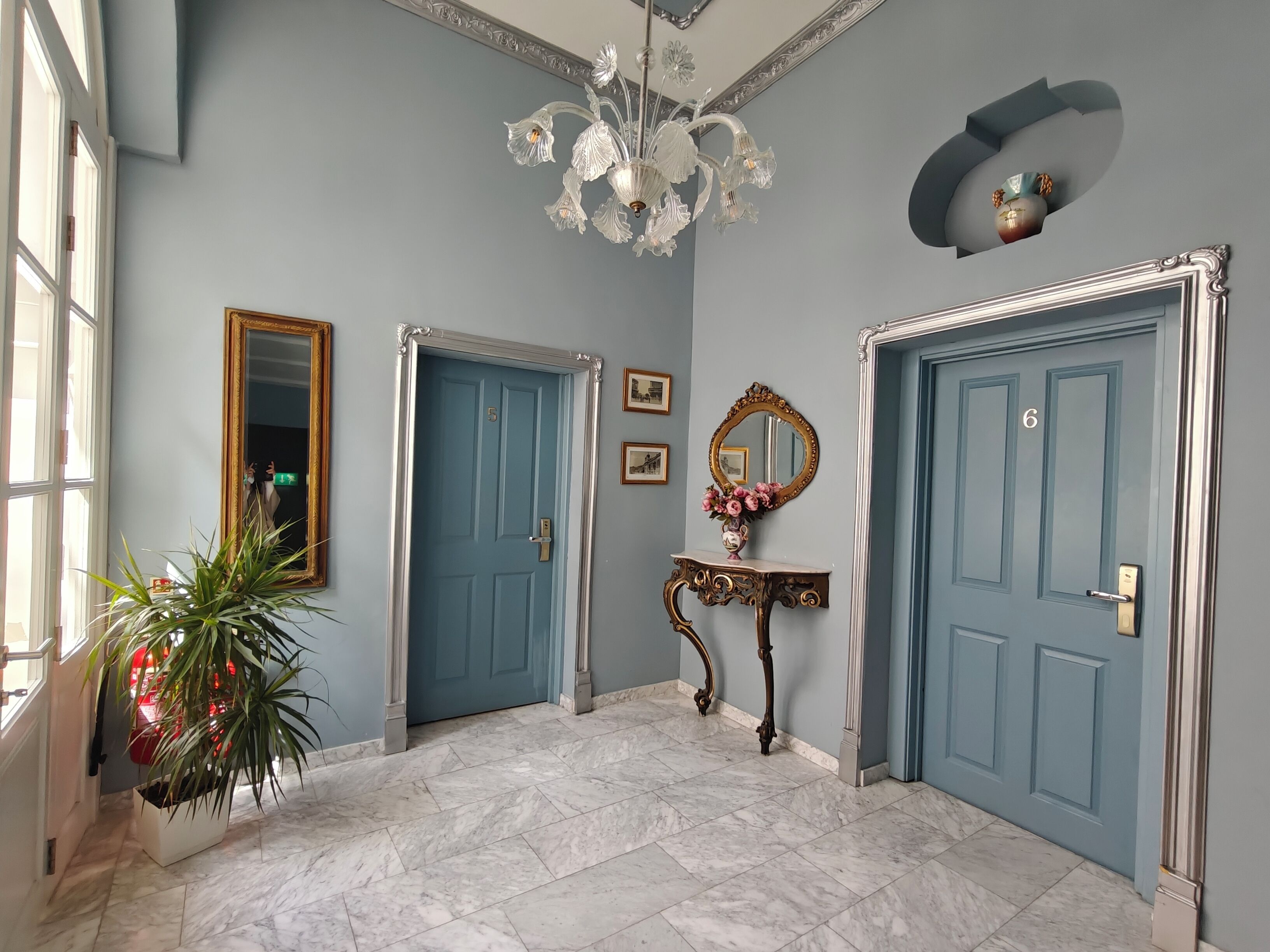 A hallway with two blue doors, a chandelier, a mirror, and a potted plant.