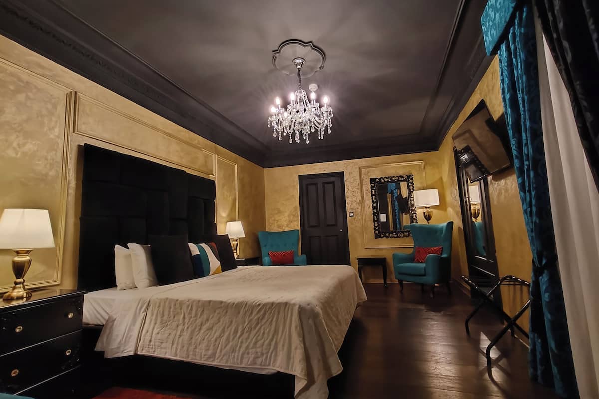 A hotel room with a large bed, a chandelier, a mirror, and a blue armchair.