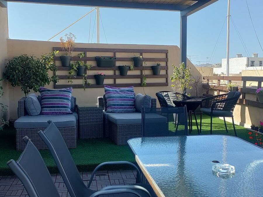 A rooftop terrace with wicker furniture, a glass table, and potted plants.