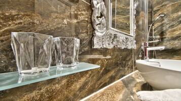 A bathroom with a marble wall, a glass shelf with two clear glasses, a white sink, and a shower area.