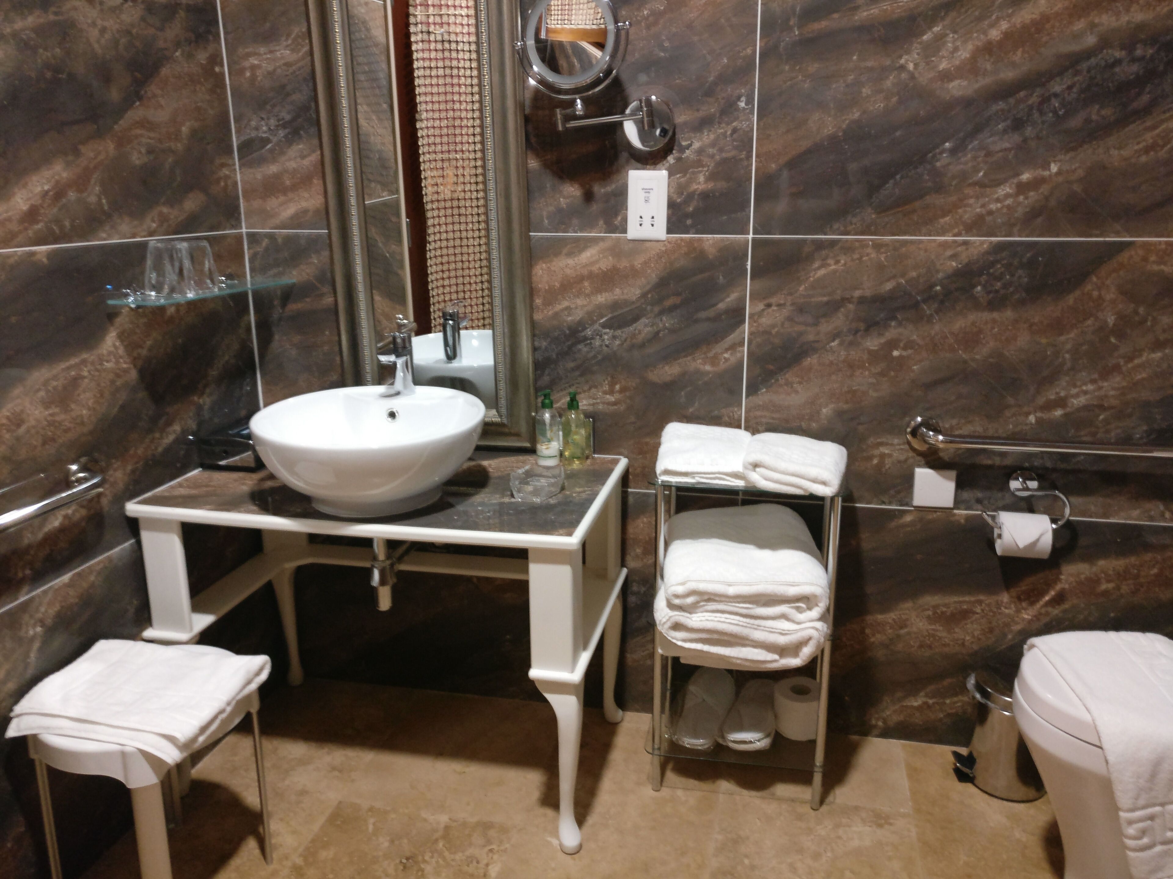 A bathroom with a white sink, mirror, and towel rack. There is a small white stool and a glass shower enclosure.