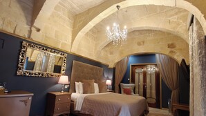 A bedroom with a stone arch, a chandelier, a large mirror, a bed with a red throw, and a wooden dresser. - Julina Boutique Living (Mosta)