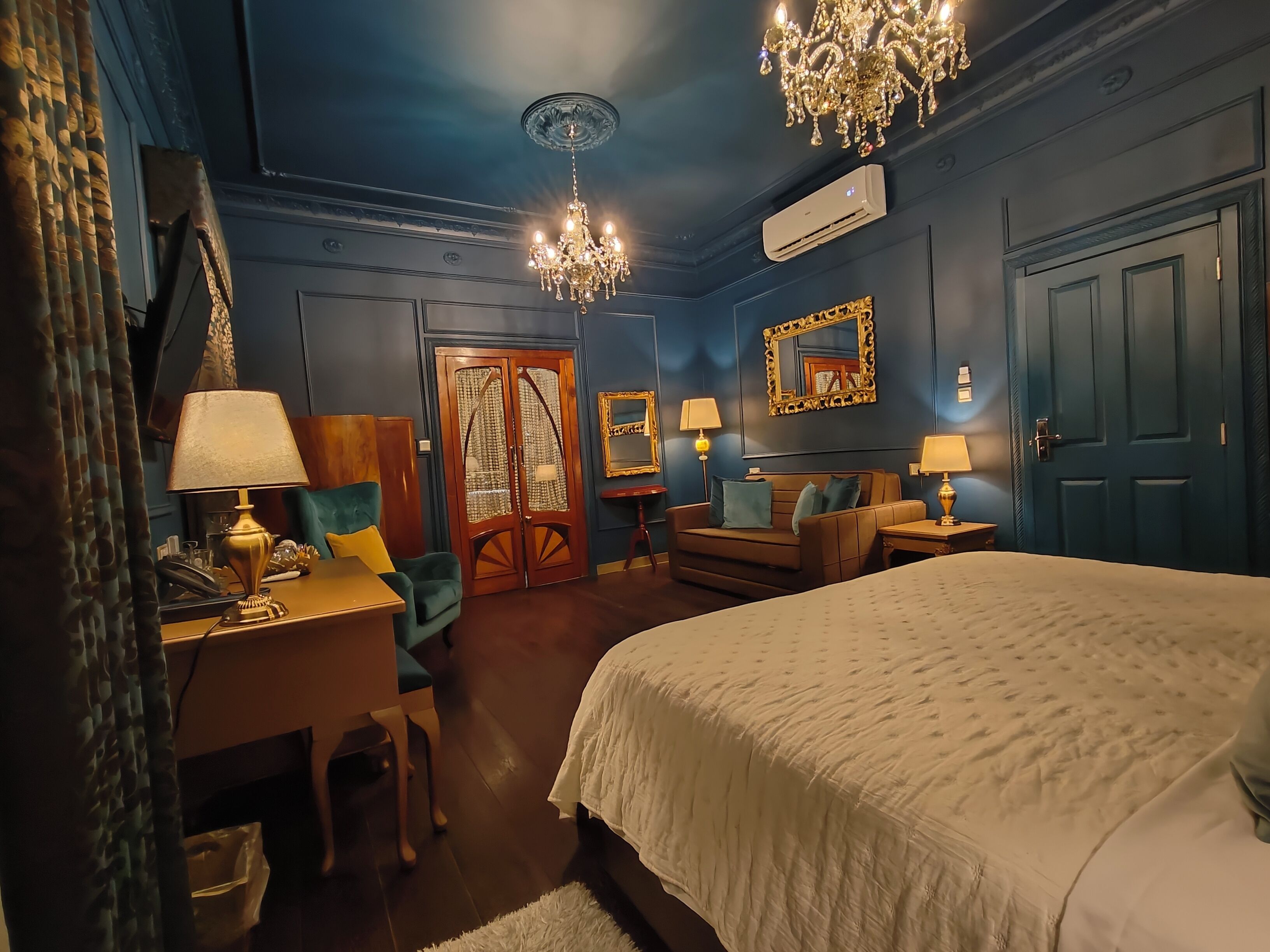 A hotel room with a bed, a desk, a chair, a dresser, a chandelier, and a mirror.