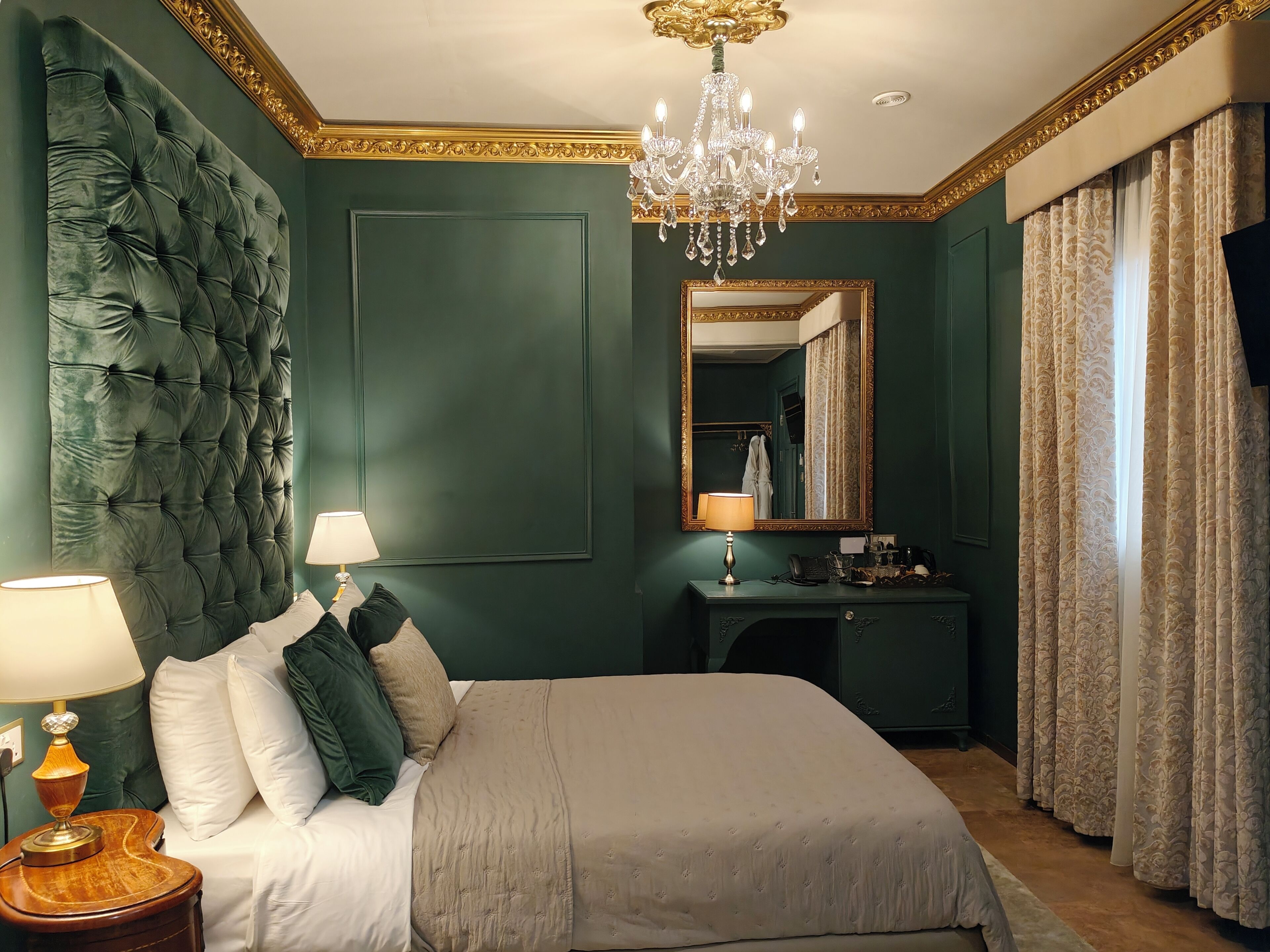 A bedroom with a tufted headboard, a chandelier, a bedside table with a lamp, a dresser, and a mirror.