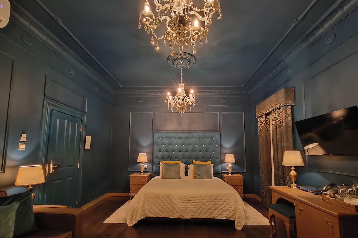 A bedroom with a large bed, two bedside tables, a chandelier, a TV, and a door.