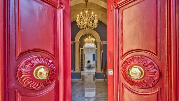 A red door with a gold knob and a chandelier above. - Julina Boutique Living (Mosta)
