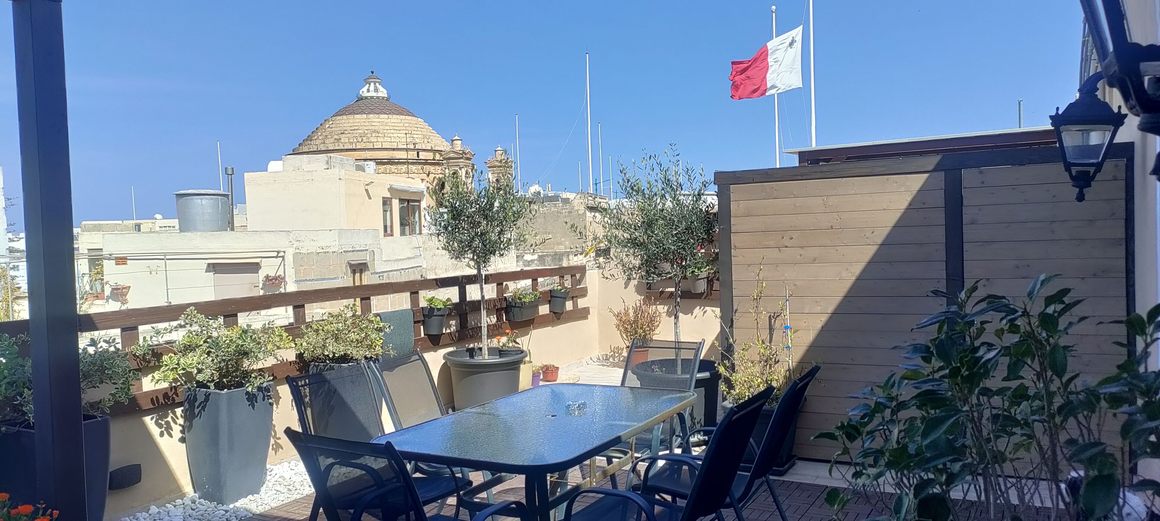 Een terras buiten met een tafel, stoelen, plantenbakken en een vlag.