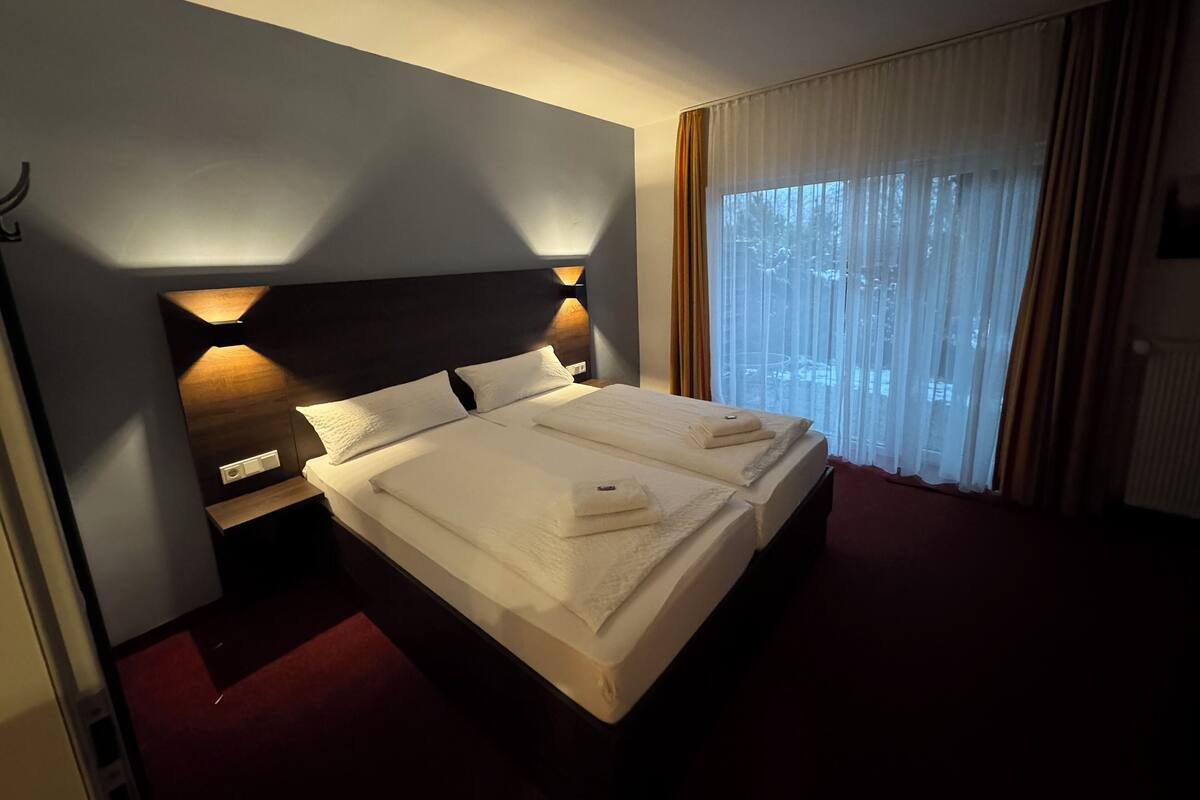 A hotel room with a large bed, a nightstand, a wall-mounted light, and a window with curtains.