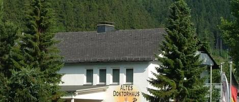 A building with a sign reading 'ALTES DOKTORHAUS' surrounded by trees and a mountain in the background.