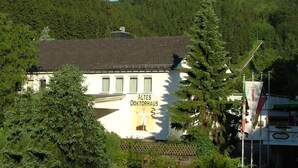 A building labeled "Altes Doktorhaus" with a white facade, surrounded by lush greenery and trees. - Altes Doktorhaus (Willingen)