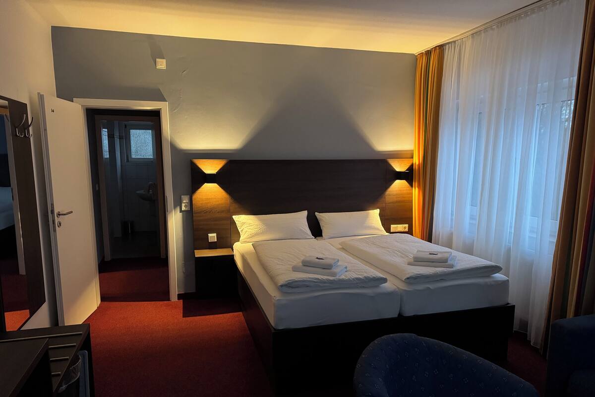 A hotel room with two beds, a desk, a chair, and a bathroom visible through an open door.