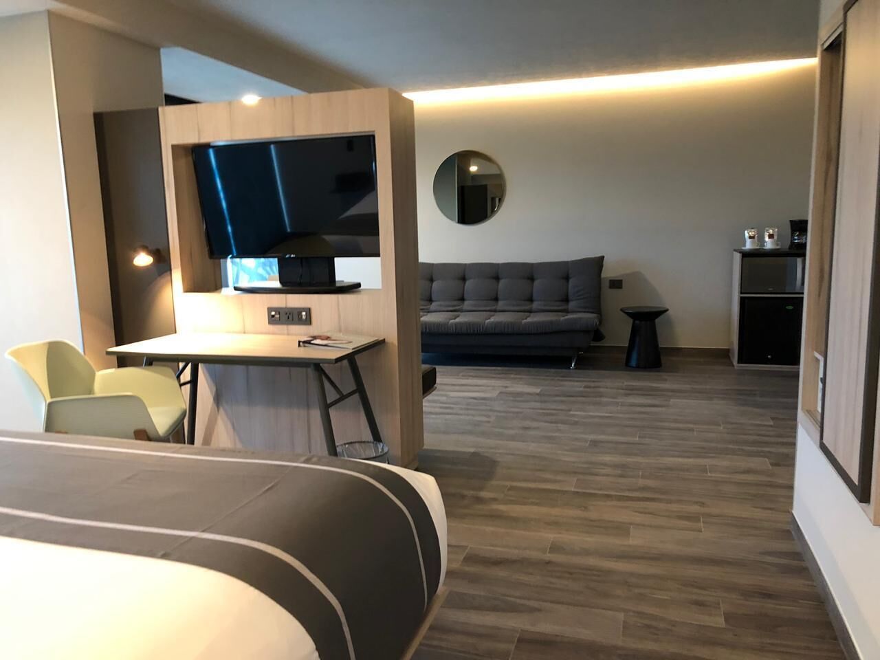 A modern hotel room with a flat-screen TV, a sofa, a dining table, and a small kitchen area.