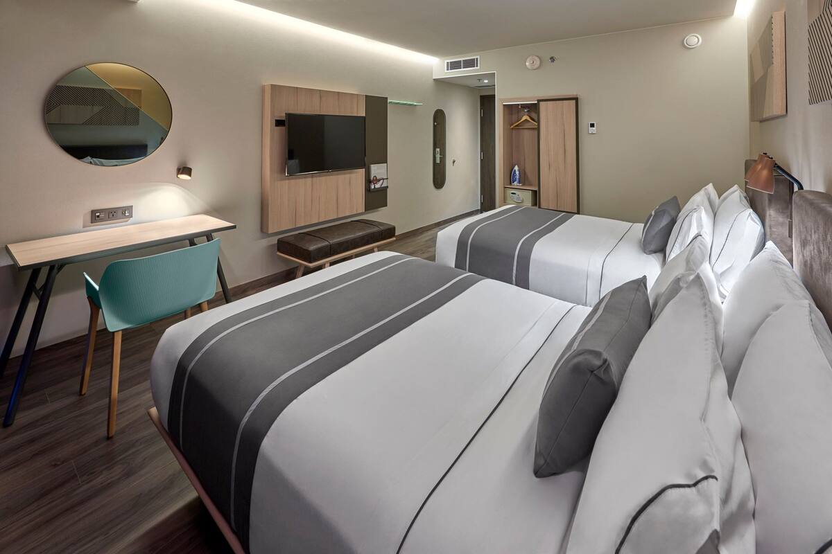 A hotel room with two beds, a desk, a chair, a mirror, a TV, and a wardrobe.