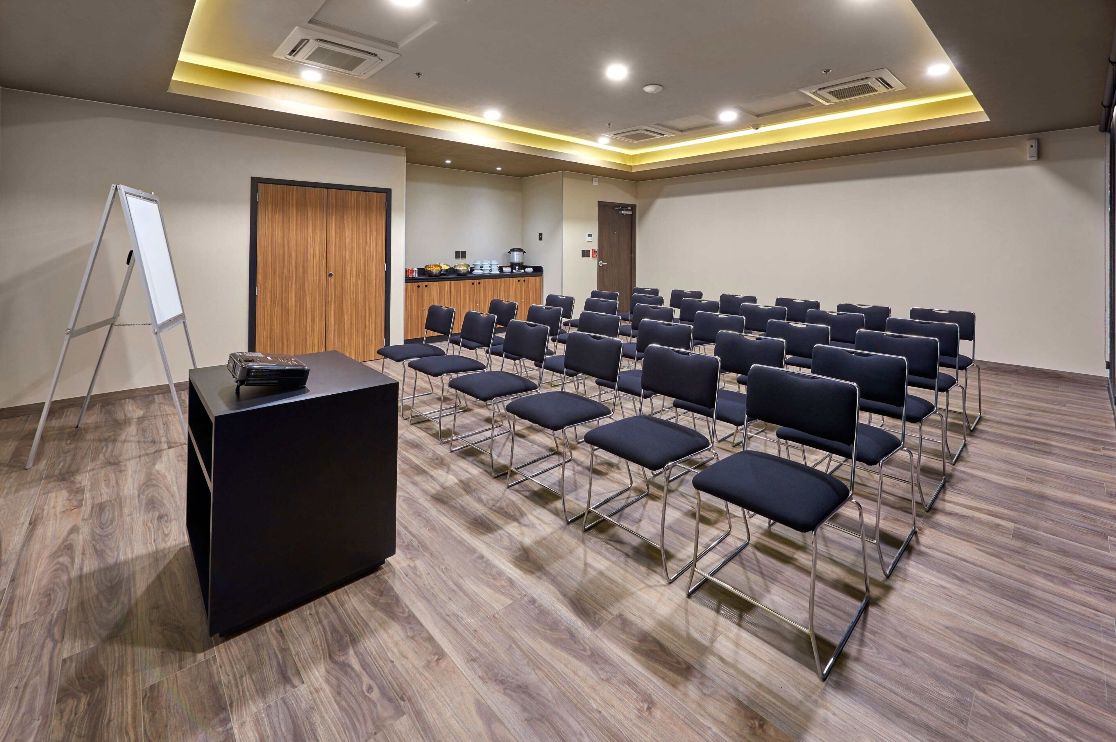 A conference room with rows of chairs, a podium, a whiteboard, and a kitchenette area.