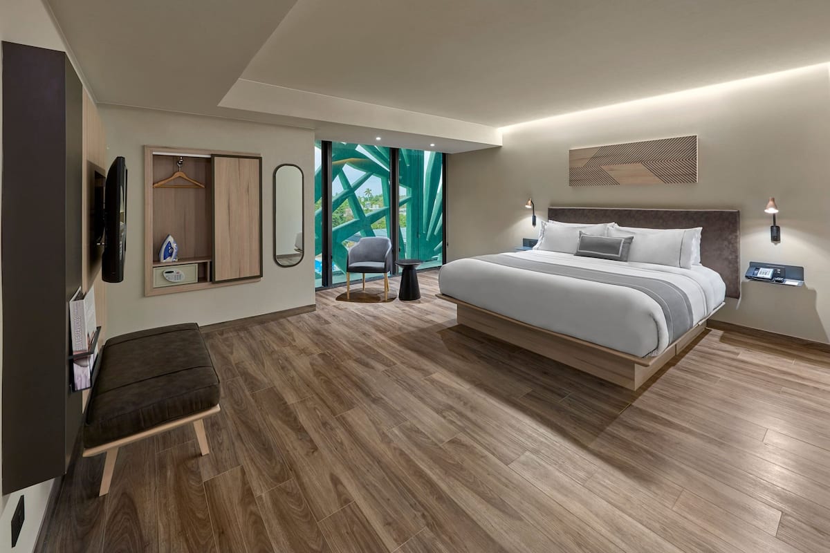 A modern hotel room with a large bed, a seating area, a TV, a wardrobe, and a view of greenery through the door.