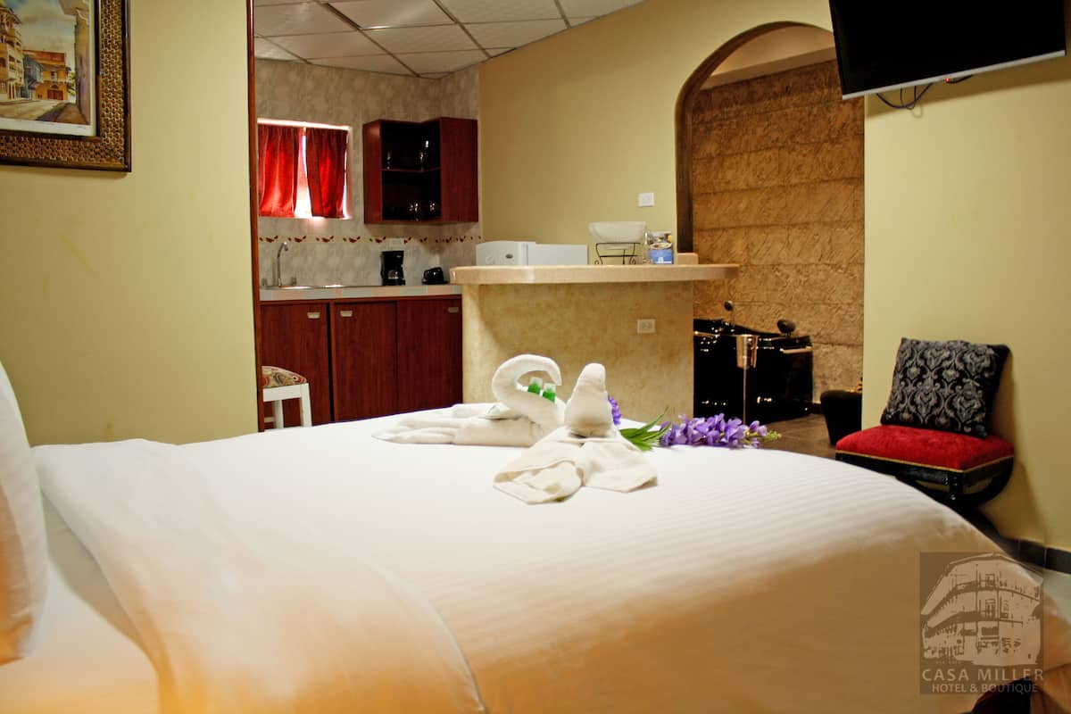 A hotel room with a bed, a small sitting area with a red cushion, a television mounted on the wall, and a kitchenette in the background.