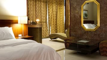 Habitación de hotel con cama, escritorio con lámpara, silla, mesita pequeña y un espejo grande en la pared.