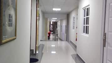 Un pasillo largo y estrecho con suelo de baldosas, paredes blancas y techo con luces fluorescentes. Hay puertas a ambos lados y un cuadro enmarcado en la pared izquierda.