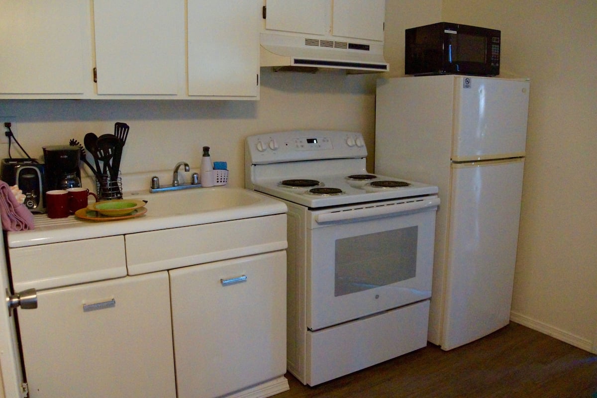 Una cocina compacta con estufa blanca, microondas y refrigerador. Hay armarios sobre la estufa y una pequeña encimera con algunos objetos.