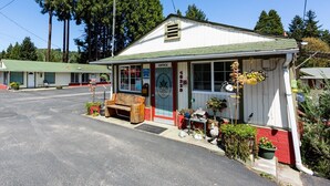 A small, single-story building with a green roof and a white exterior. It has a porch with a wooden bench and potted plants. An American flag is flying above the building. - Arbor Inn Motel Oakridge (Oakridge)