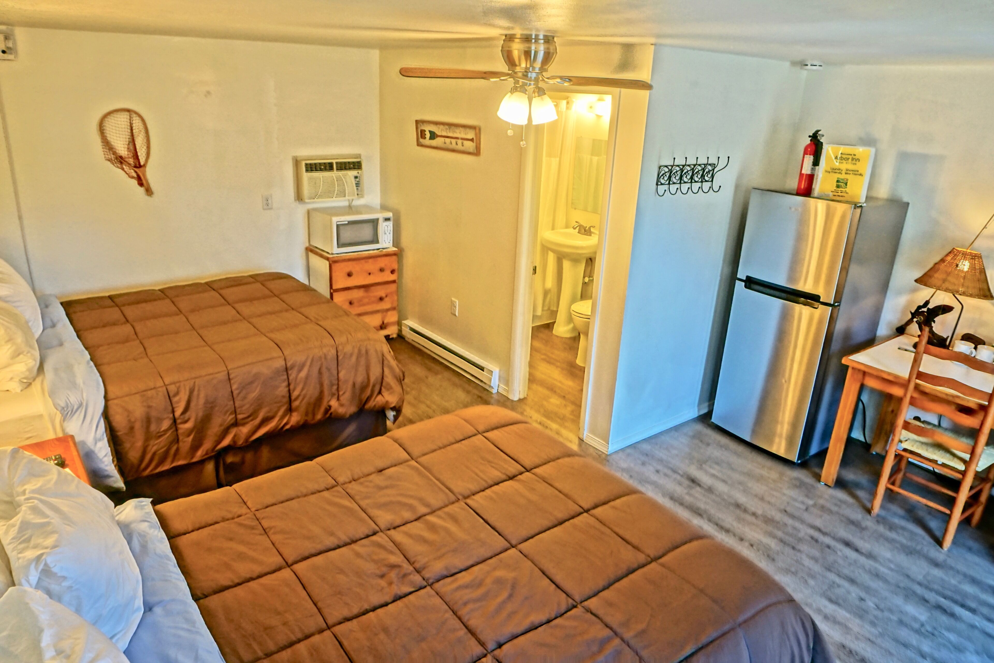 A compact hotel room with two beds, a kitchenette, a ceiling fan, and a bathroom visible through an open door.