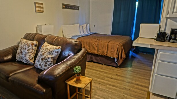 A hotel room with a brown leather sofa, a small wooden side table with a plant, a bed with a brown cover, a white desk with a coffee maker, and a window with blue curtains. - Arbor Inn Motel Oakridge (Oakridge)