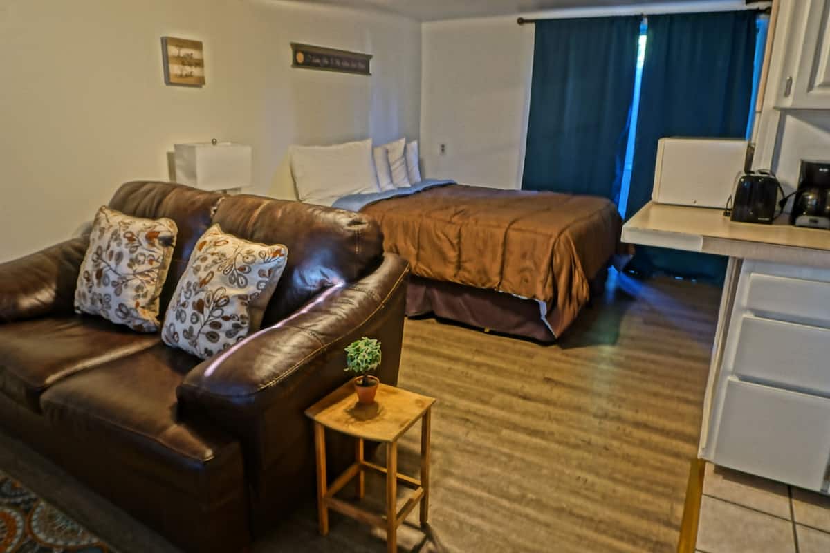 Habitación de hotel con un sofá de cuero marrón, una mesita de noche de madera con una planta, una cama con cubrecama marrón, un escritorio blanco con cafetera y una ventana con cortinas azules.