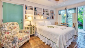 Un dormitorio con cama, un sillón floral, una estantería y vistas al verdor por la ventana.