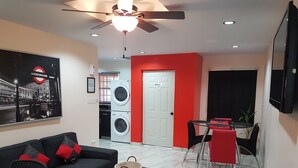 Un salon moderne comprenant un canapé, une table basse, un téléviseur, un ventilateur de plafond, un lave-linge et un coin repas avec des chaises et une table.