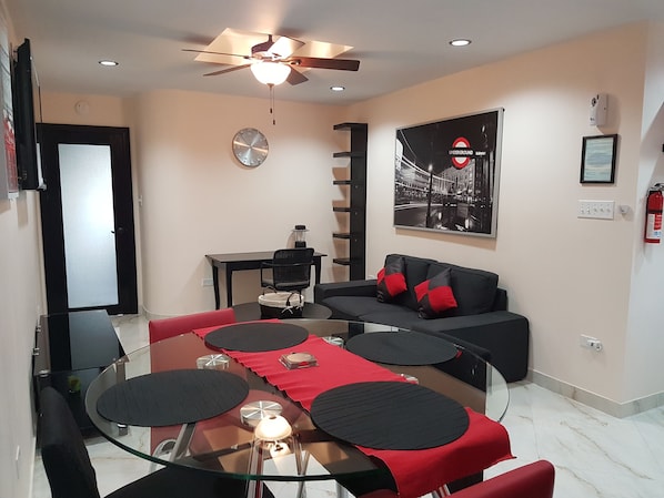 A modern living room with a glass dining table, red and black cushions, a black sofa, a ceiling fan, a clock, and a poster of the London Underground.