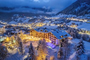 Une ville de montagne enneigée, illuminée la nuit, avec des bâtiments éclairés et un hôtel imposant, doté de plusieurs étages et de balcons.