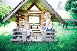 Deux enfants sont assis sur un banc en bois devant une construction rustique également en bois, dotée d’un toit en pente.
