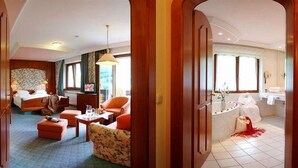 Hotelkamer met een bed, zithoek met een bank en fauteuils, een tafeltje met een lamp, en een badkamer met bad en wastafel.