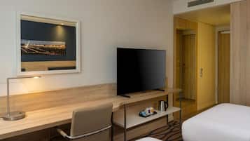 A hotel room with a flat-screen TV, a desk with a chair, a bed, and a framed picture on the wall.