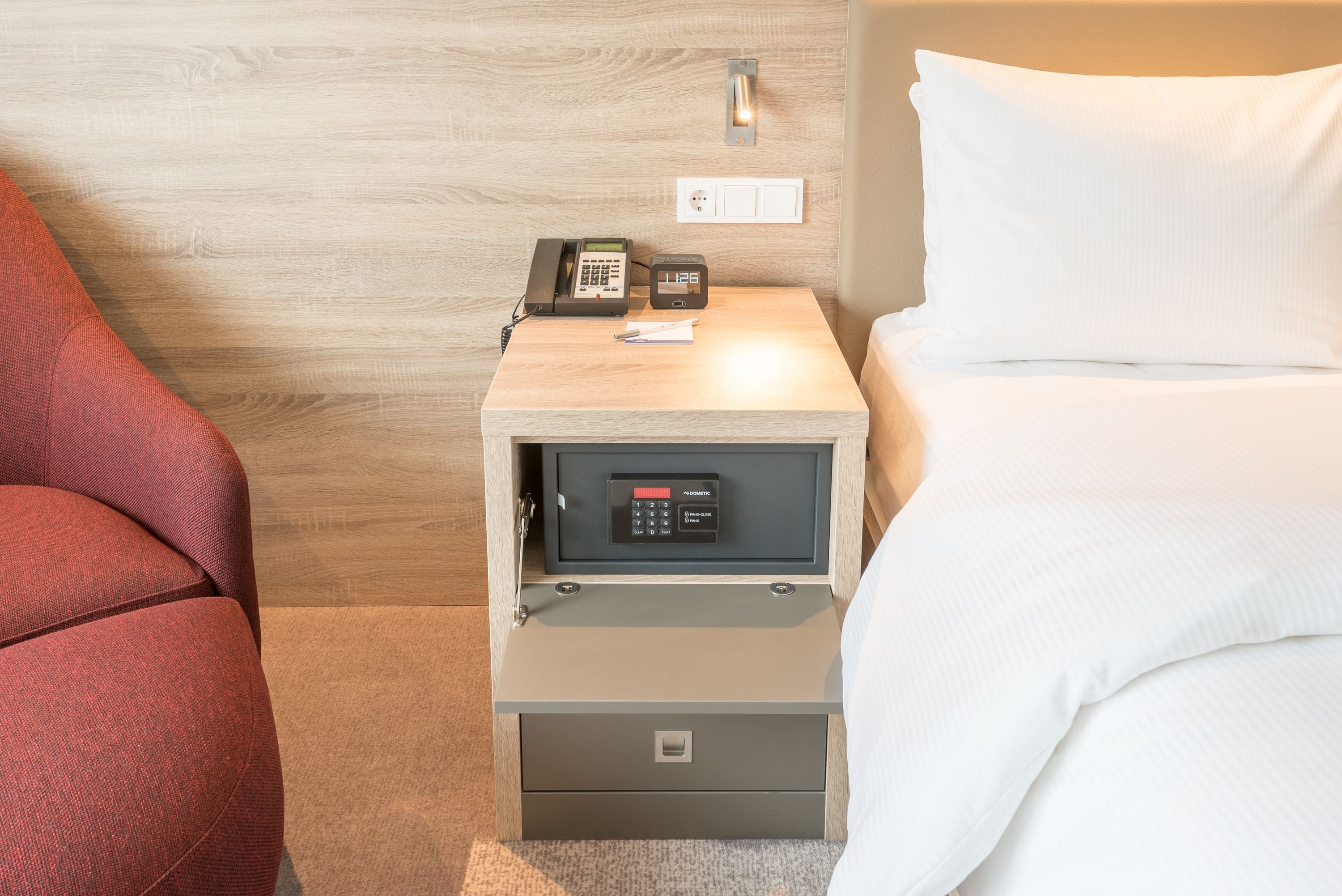 Habitación de hotel con pared de madera, mesita de noche con teléfono, caja fuerte y cama con ropa blanca.
