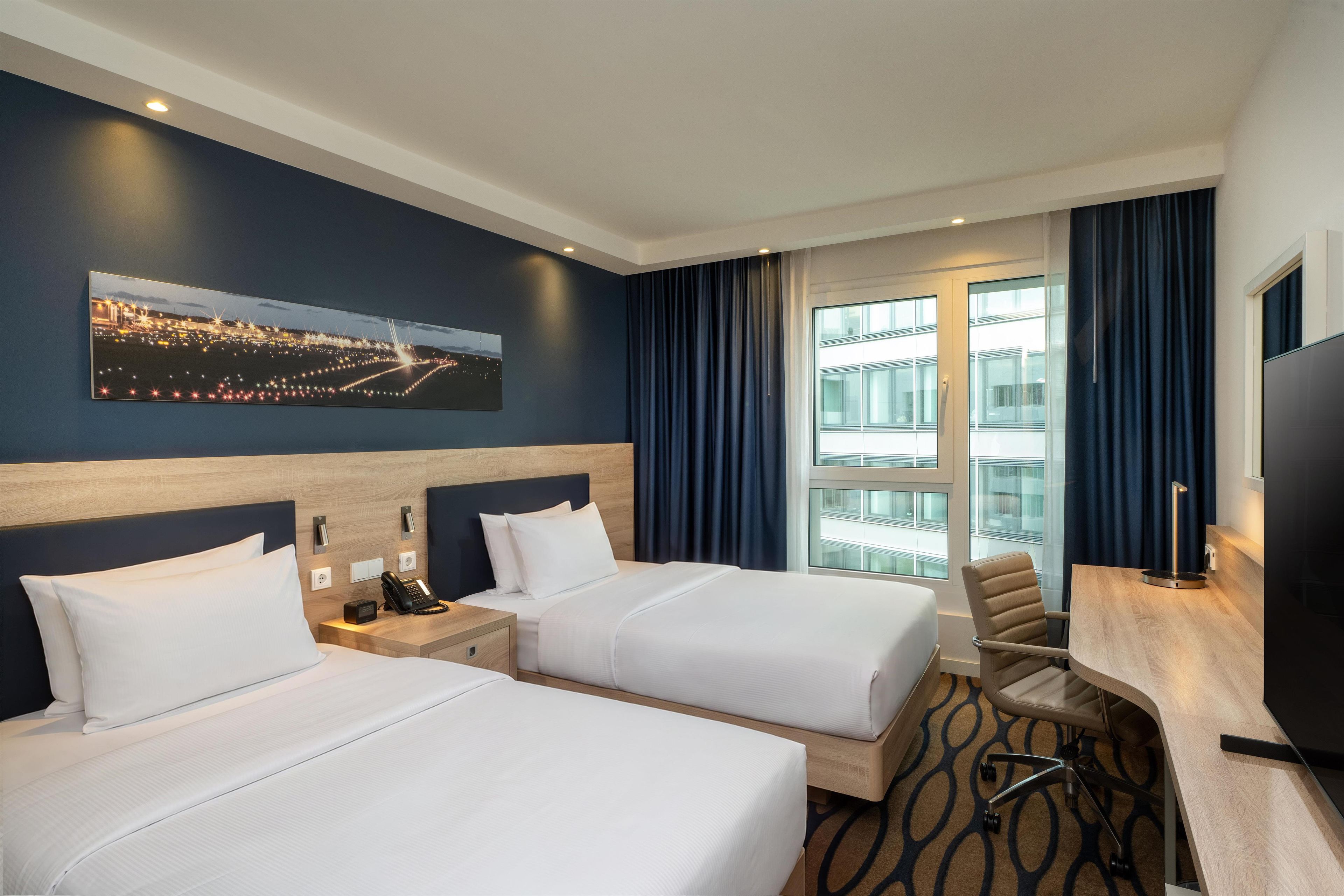 A hotel room with two beds, a desk, a chair, a flat-screen TV, and a cityscape painting on the wall.